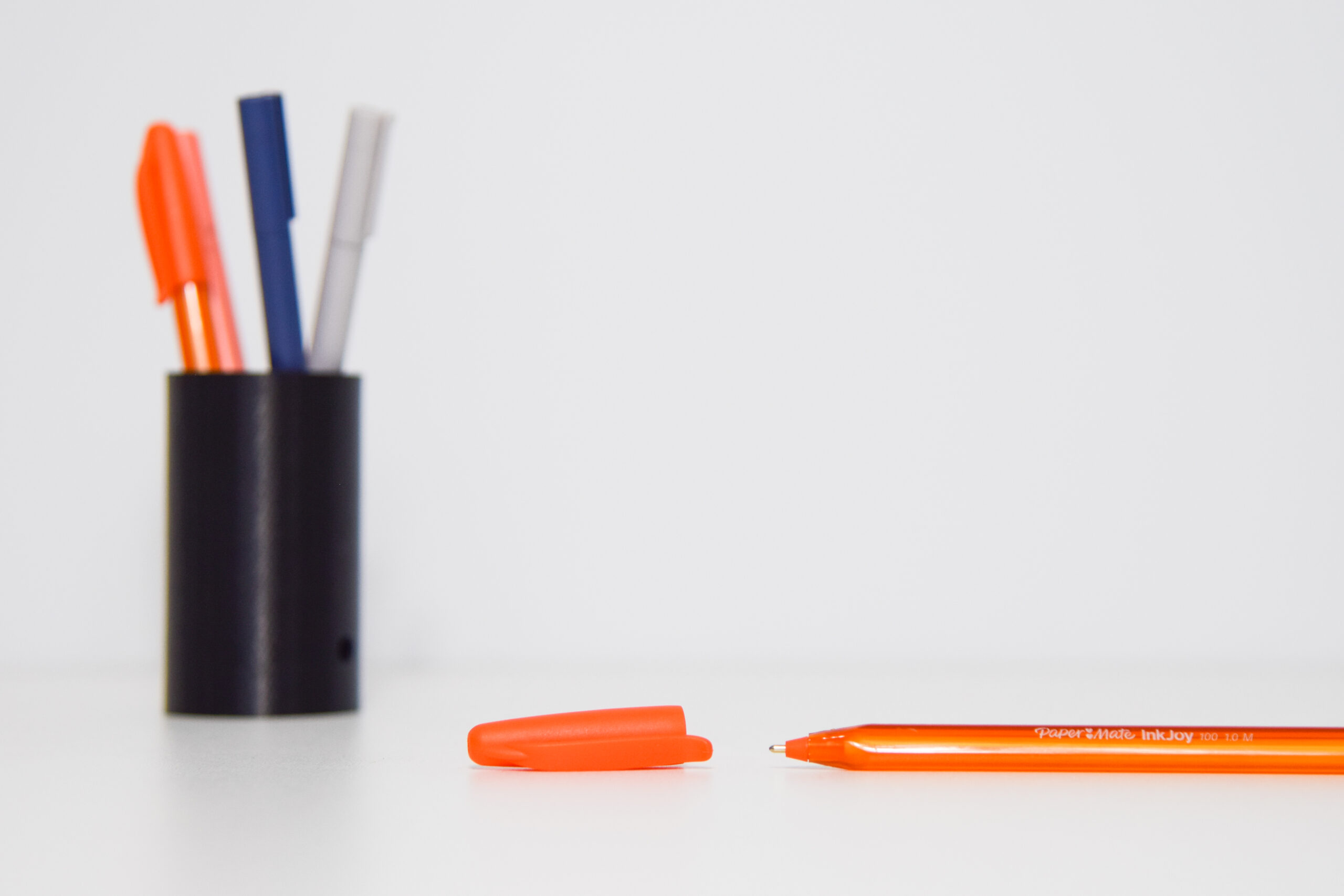 A black pen holder holds 4 pens (2 orange, 1 navy and 1 grey), and an orange pen lays with its cap off in front of it.